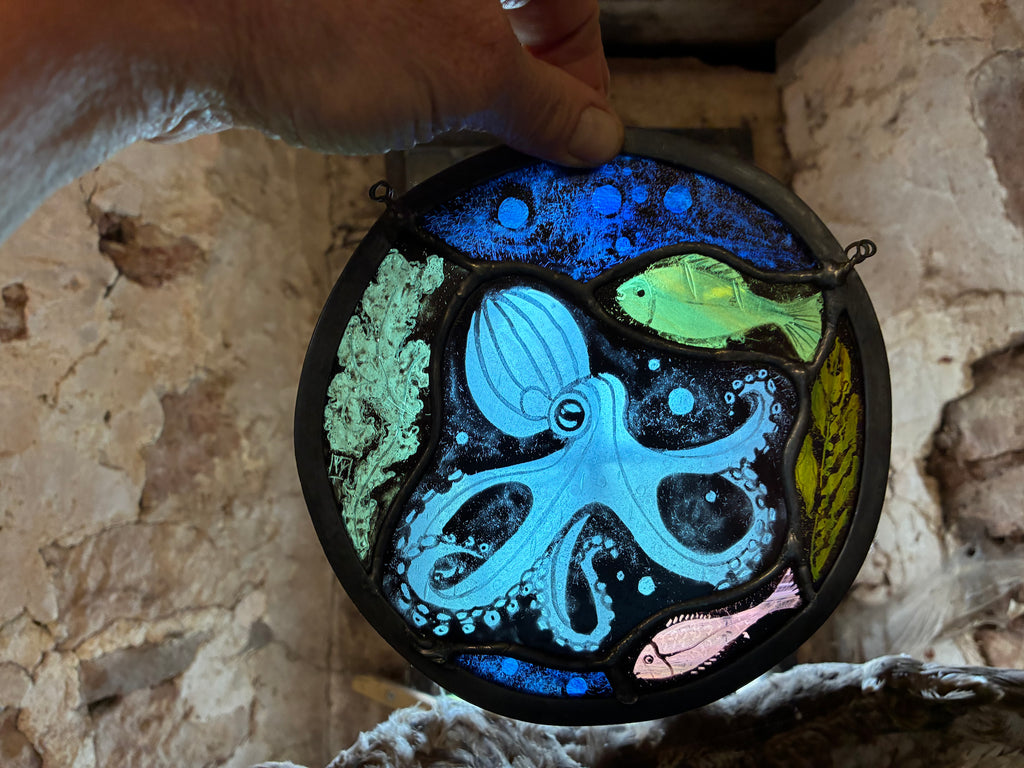 A hand holds a round stained glass suncatcher featuring an underwater scene with a light blue octopus, green fish, pink fish, and coral against a dark blue and black background, set against a rustic stone wall.