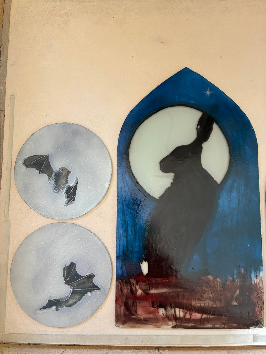 Hand-painted stained glass panels featuring a hare silhouette against a full moon in a blue arched window shape, alongside two round glass pieces with bat silhouettes, displayed on an unlit light box.