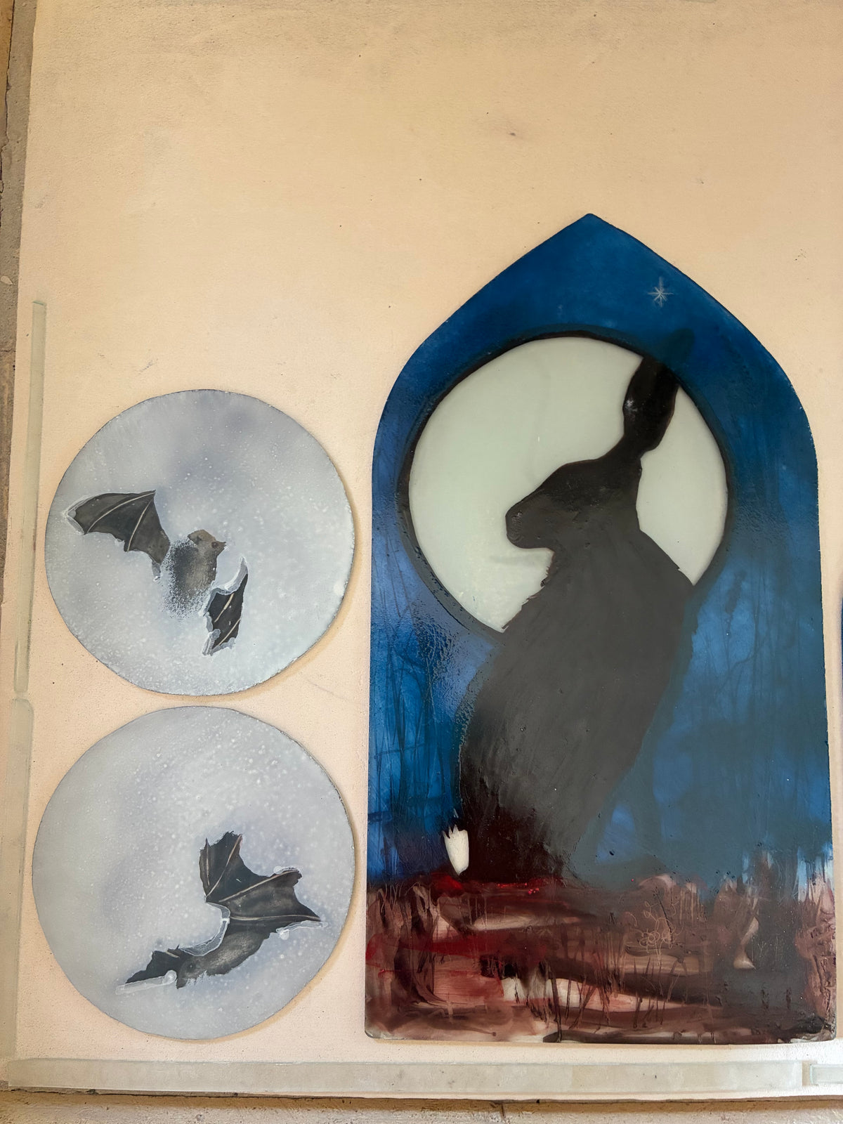 Hand-painted stained glass panels featuring a hare silhouette against a full moon in a blue arched window shape, alongside two round glass pieces with bat silhouettes, displayed on an unlit light box.