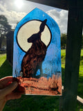 A hand holds a stained glass art piece depicting a brown hare in front of a large cream moon against a textured blue sky, photographed outdoors in a grassy garden.