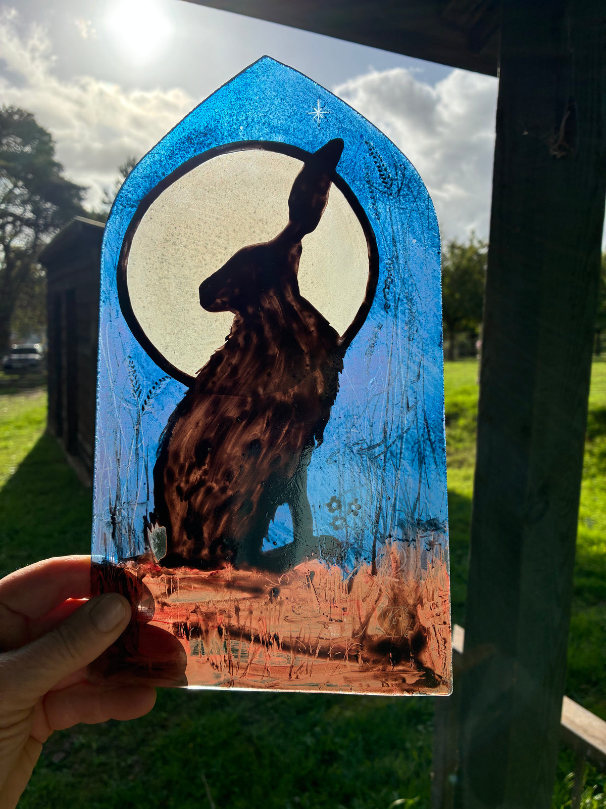 A hand holds a stained glass art piece depicting a brown hare in front of a large cream moon against a textured blue sky, photographed outdoors in a grassy garden.