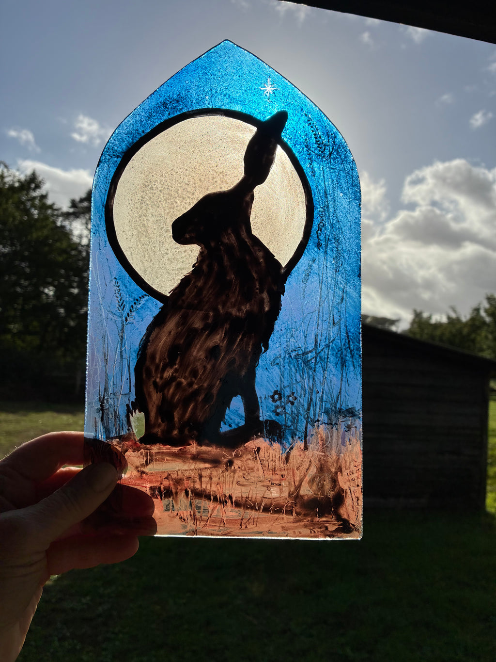 A hand holds a stained glass window panel featuring a black hare silhouette in front of a large cream moon, with a blue arched top and a brown ground, photographed outdoors against a cloudy sky and trees.
