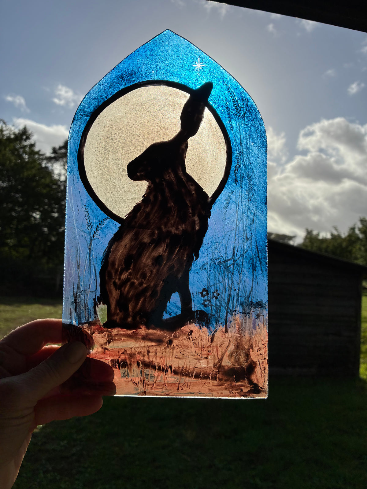 A hand holds a stained glass window panel featuring a black hare silhouette in front of a large cream moon, with a blue arched top and a brown ground, photographed outdoors against a cloudy sky and trees.