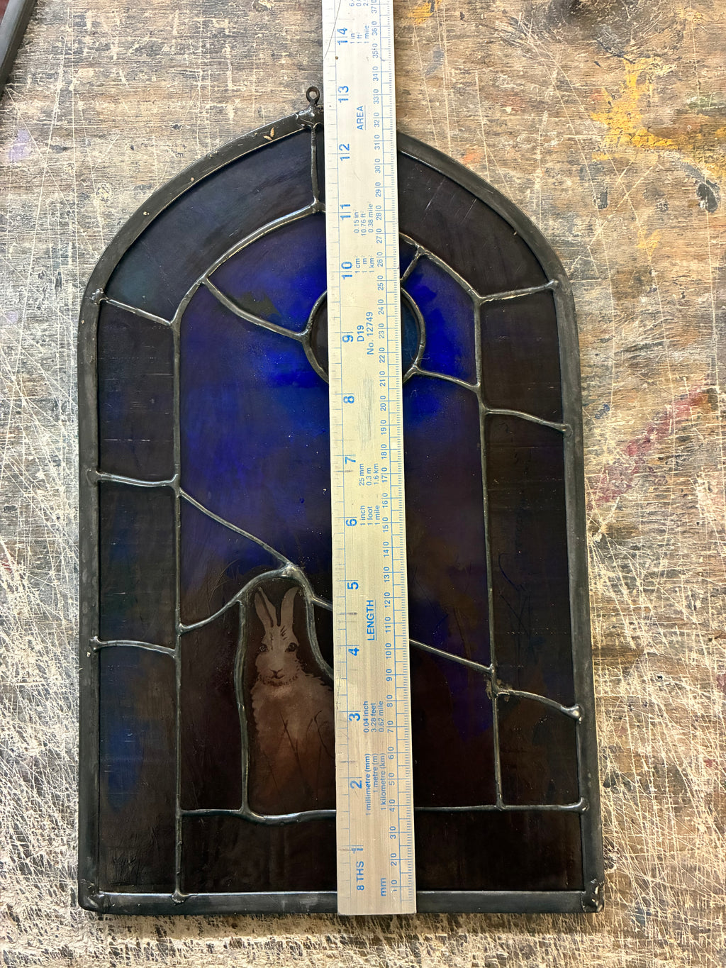 An arched stained glass window panel featuring a rabbit in the lower left and a deep blue sky motif, with dark metal leading and a wooden ruler placed vertically in the center for scale, set against a scratched wooden workbench background.