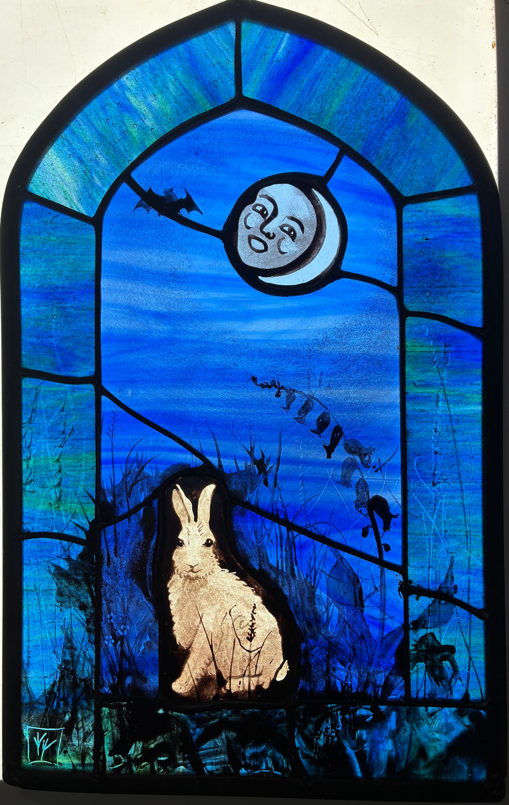 A stained glass artwork depicting a white hare in a dark field beneath a crescent moon.  A bat is flitting in the dark blue sky and the whole is framed by a green-teal arched border.