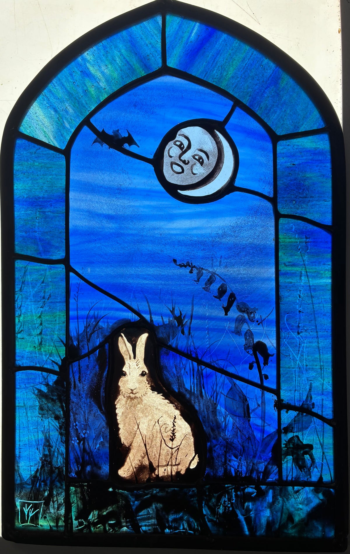 A stained glass artwork depicting a white hare in a dark field beneath a crescent moon.  A bat is flitting in the dark blue sky and the whole is framed by a green-teal arched border.