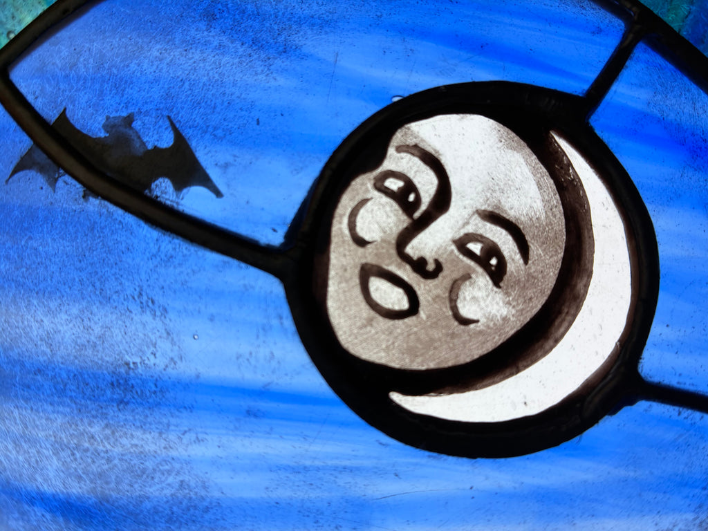 A close-up of a stained glass panel featuring a stylized new moon face with a bat silhouette against a textured blue background.