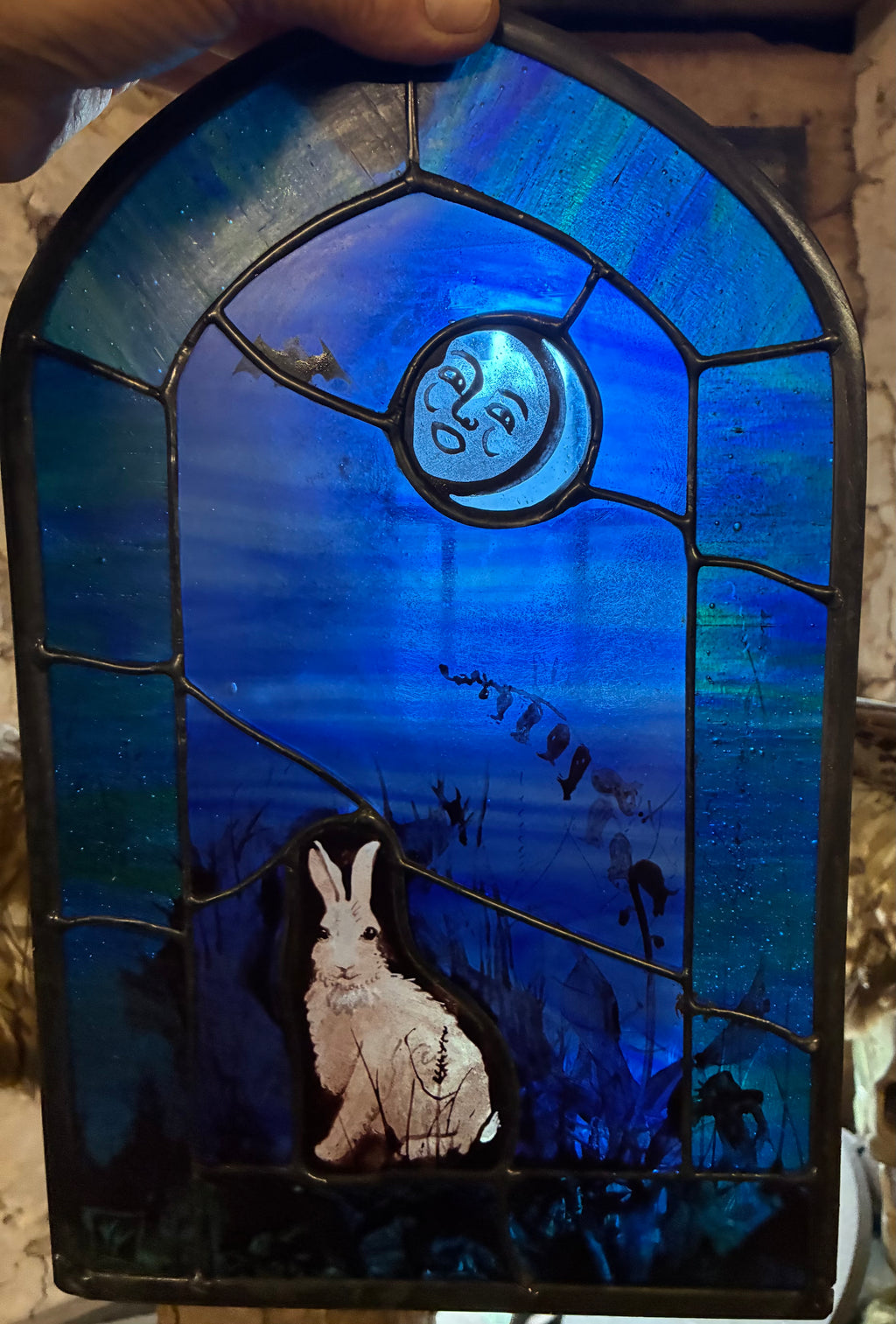 Handheld stained glass art piece featuring a blue night sky with a crescent moon and a white rabbit, framed in dark metal.
