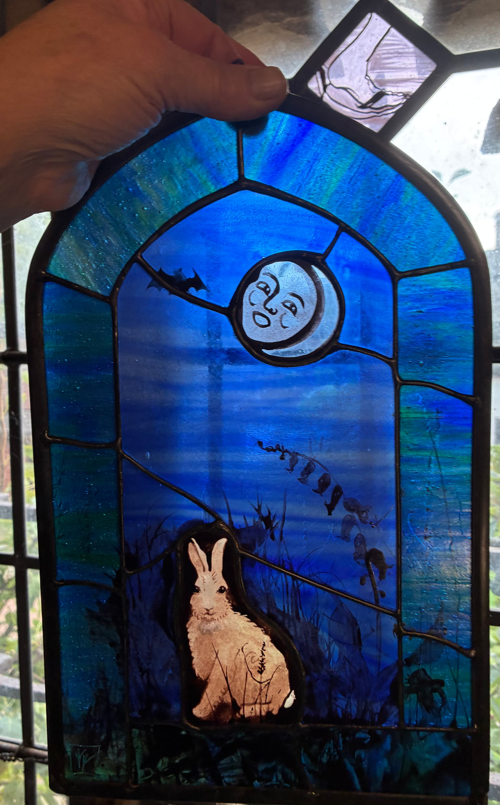 A hand holds a stained glass panel featuring a blue night sky with a moon, a bat, and a rabbit, framed by black solder lines.