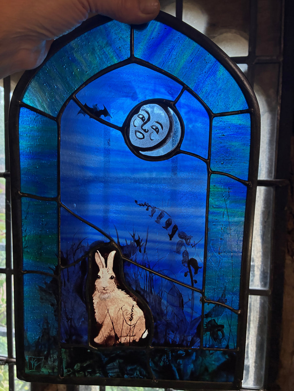 Hand-held stained glass window panel featuring a blue night sky with the new moon, a bat, a rabbit, and a foxglove silhouette, framed in dark metal.
