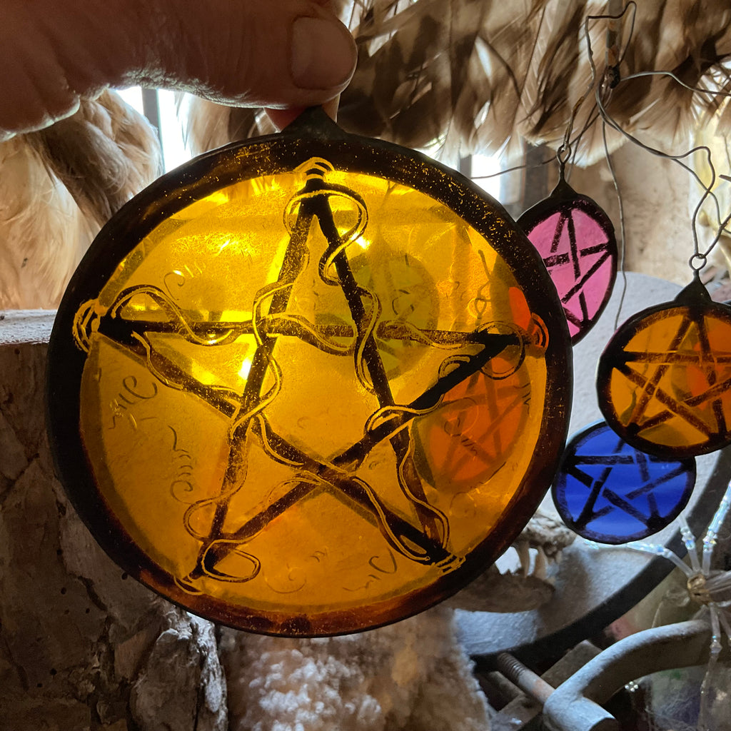 Decorative stained glass pentacle ornament with a five star design in a circle held by a hand against a blurred background.  Smaller pentacles are shown in the background.