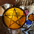 Decorative stained glass pentacle ornament with a five star design in a circle held by a hand against a blurred background.  Smaller pentacles are shown in the background.