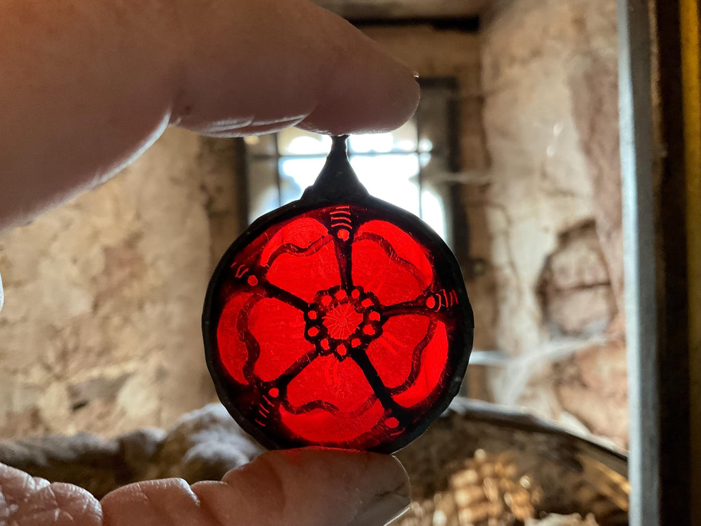 Red and black stained glass Tudor rose held by a hand in front of a stone wall with a leaded window