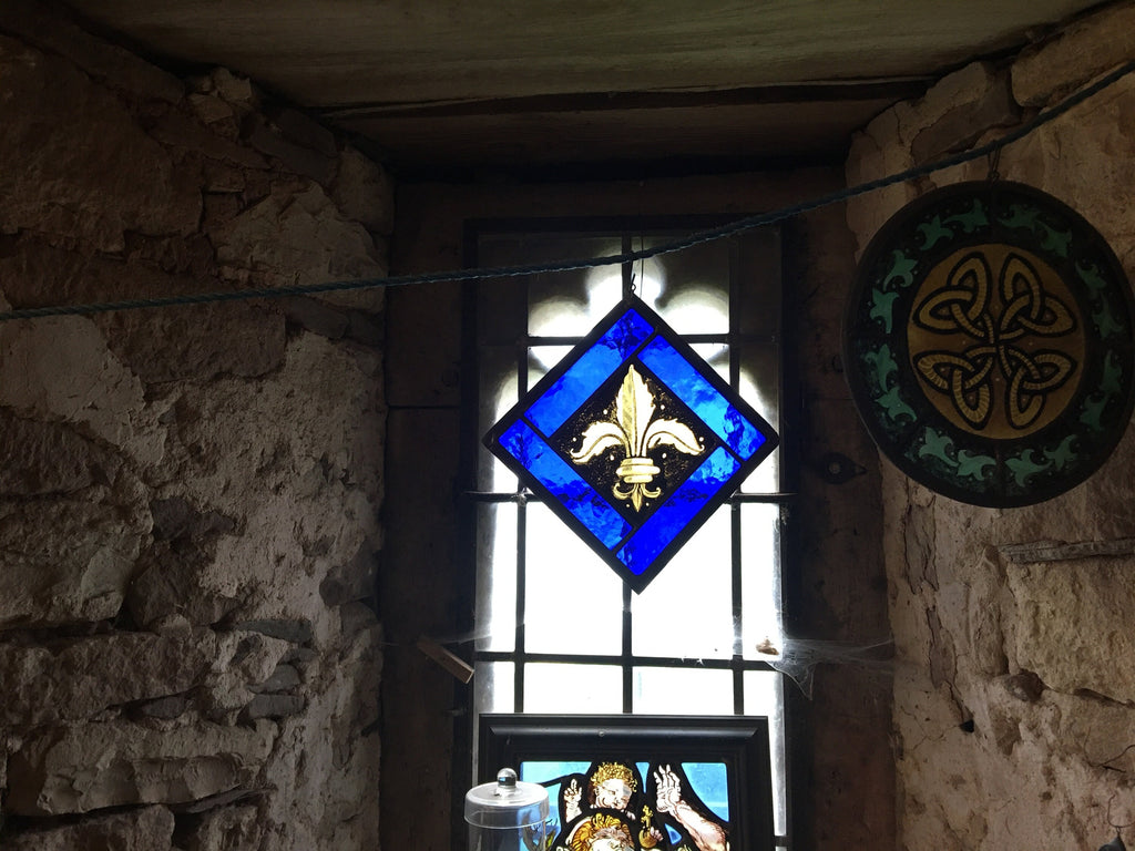 Stained glass window with a blue diamond design featuring a fleur-de-lis, in a stone-walled room.