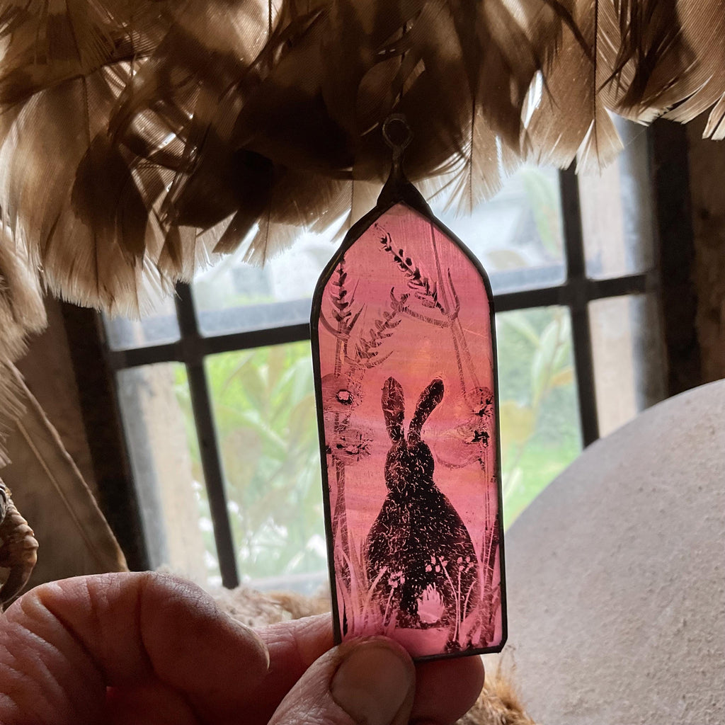 Decorative pink pendant with a rabbit design held by a hand, with feathers and a blurred background.
