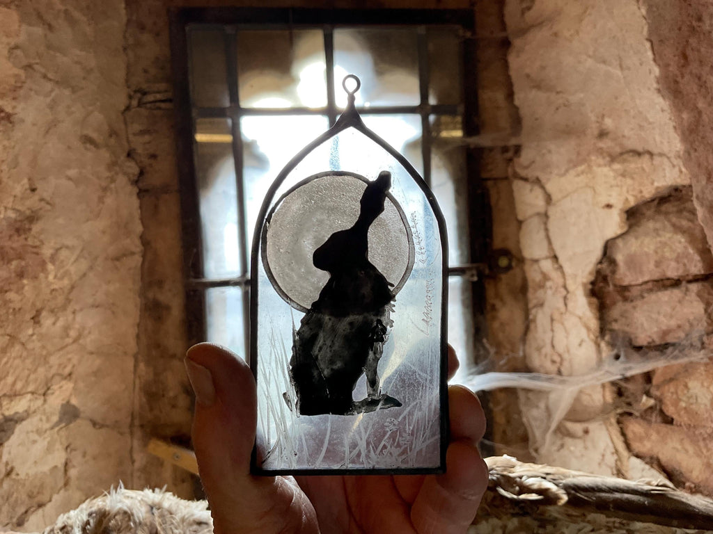 Hand holding a decorative glass panel with a silhouette of a hare and a full moon against a stone wall background